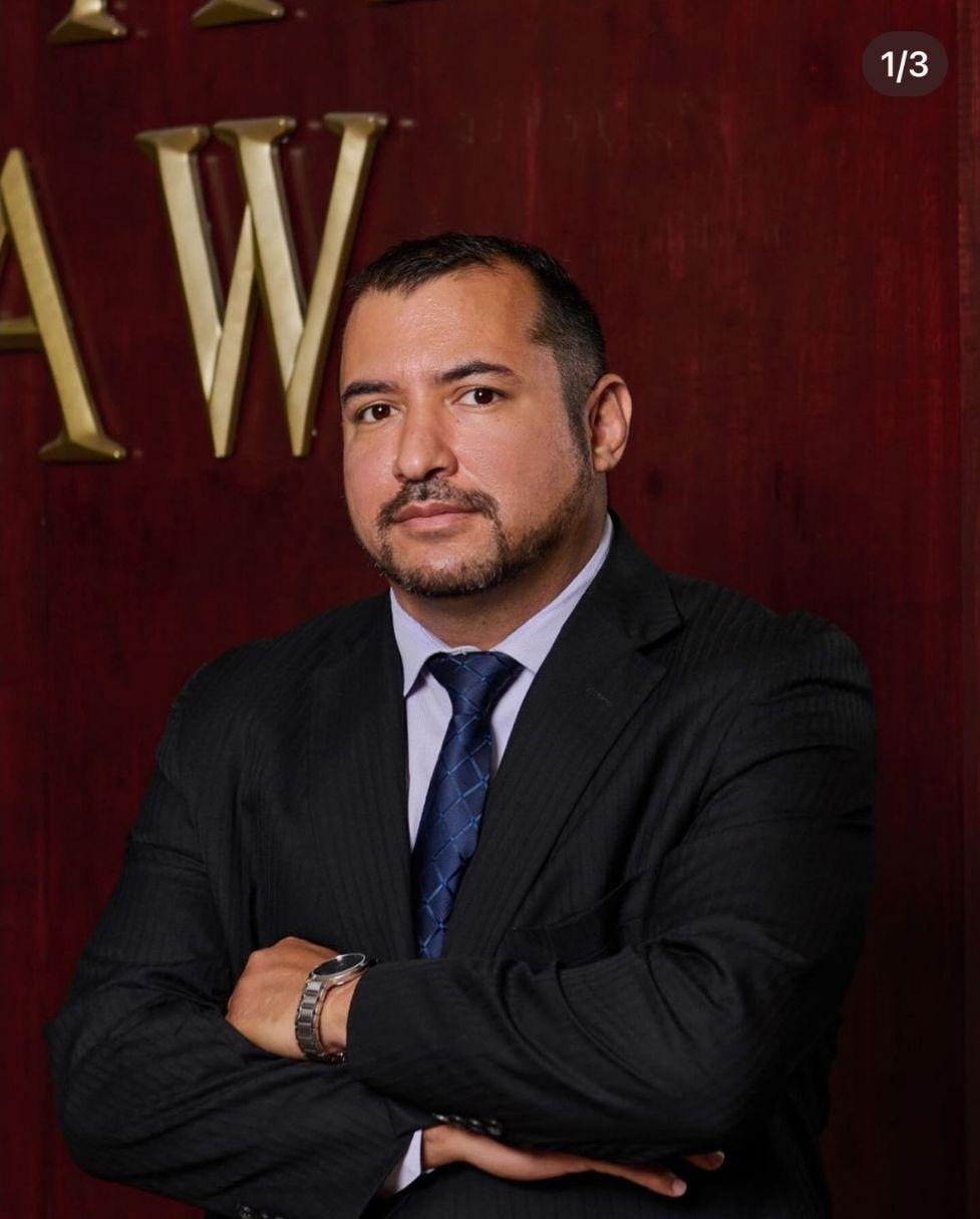 Man in a suit with arms crossed in front of a reddish-brown background with gold letters.